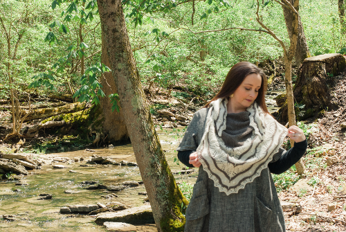 Mindy is standing in front of a small stream in a wooded area. She is looking down at the lace shawl she is wearing.